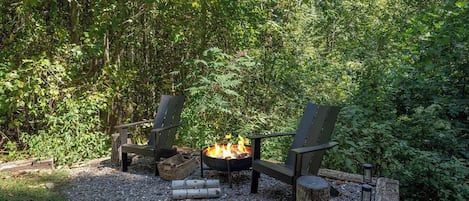 A fire pit with two Adirondack chairs and a log fire in a forested area.