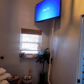 A room with a television displaying 'Roku TV,' a window with blinds, a plant, a small shelf with items, and a wall-mounted telephone. - Boho Mtn Vibe'z~ Hot Tub+Fire Pit~ 3 mins to BRP! (Spruce Pine)