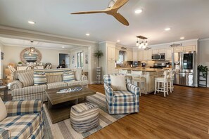 A spacious living room with a sofa, armchair, ottoman, and a coffee table. There is a kitchen area with a refrigerator, microwave, and bar stools.