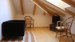 Een kleine zolderkamer met een schuine kap, houten balken, een zwarte bank, twee rieten stoelen, een televisie en een geweven mand.