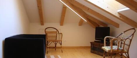 A small attic room with a sloped ceiling, wooden beams, a black sofa, two wicker chairs, a television, and a woven basket.