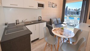 A compact kitchen with a microwave, sink, and kettle, a dining area with a table and chairs, and a view of a pool and buildings outside. - Holiday Home Near Bukowo Lake and Beaches (Darlowo)