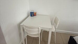 Una pequeña mesa blanca con dos sillas en un rincón de una habitación de paredes blancas y suelo de baldosas.