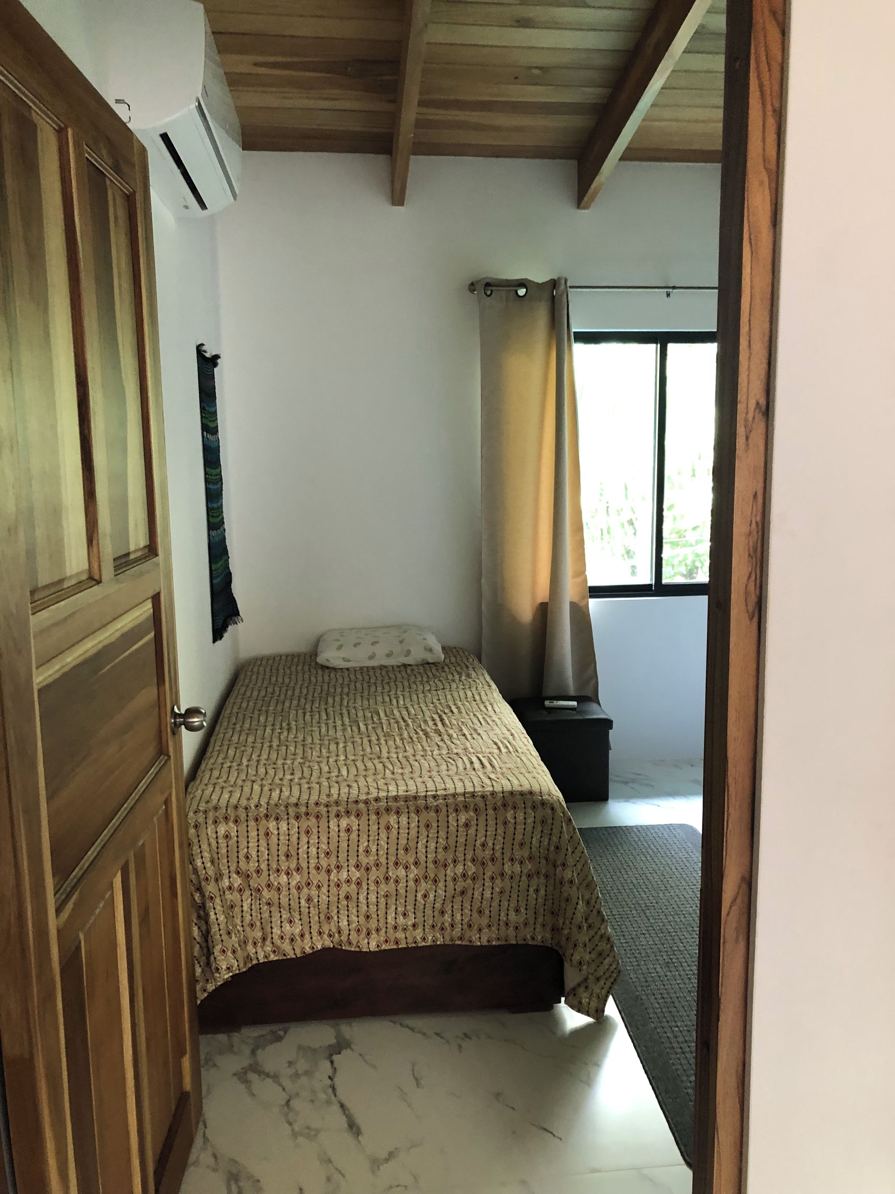 Un dormitorio con cama, puerta de madera, aire acondicionado, ventana con cortinas y una mesita de noche pequeña.