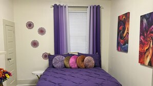 A bedroom with a purple bed, a bench, a white door, a small table, and a vase with flowers.