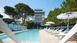 A swimming pool with lounge chairs and umbrellas, surrounded by trees and a multi-story building. - Apartment in Pietra Ligure With Garden (Pietra Ligure)