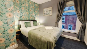 Un dormitorio con papel tapiz floral, una cama con ropa de cama verde, una mesita de noche, una ventana con cortinas y un cuadro en la pared.