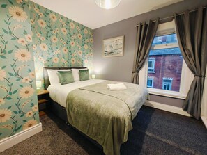 A bedroom with floral wallpaper, a bed with green bedding, a nightstand, a window with curtains, and a painting on the wall.