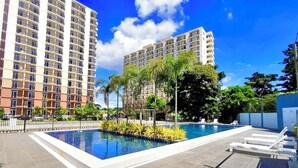 A swimming pool with lounge chairs, surrounded by palm trees and a tall building in the background. - Studio Unit W/ Balcony•Sea & City View (Lapu-Lapu)
