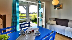A room with a dining table set for two, a bed with a gray headboard, and a view of a green landscape through an open door. - Comfortable Holiday Homes With Terraces (Postomino)