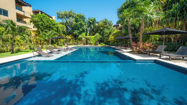 A swimming pool with lounge chairs and umbrellas, surrounded by lush greenery and a multi-story building. - Vaiven 124 - 2 Recamaras 4 Piscinas (Playa del Carmen)