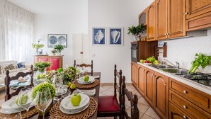Un'area pranzo con tavolo in vetro apparecchiato per quattro, circondato da sedie in legno. Si intravede a destra una cucina con pensili in legno e un lavello.