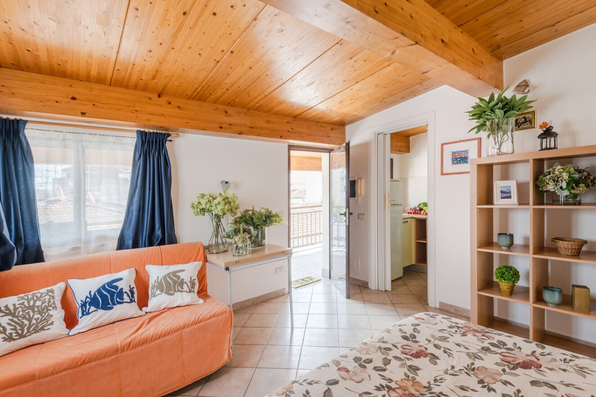 A cozy living room with a wooden ceiling, a sofa with decorative pillows, a coffee table, and a bookshelf with plants and decorative items.