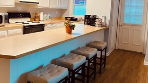 Una cocina moderna con armarios blancos, una isla central con taburetes, horno empotrado y una ventana con persianas.
