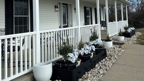 A two-story house with a covered porch, white siding, and black shutters. There are potted plants on the porch and a gravel pathway leading to the house.