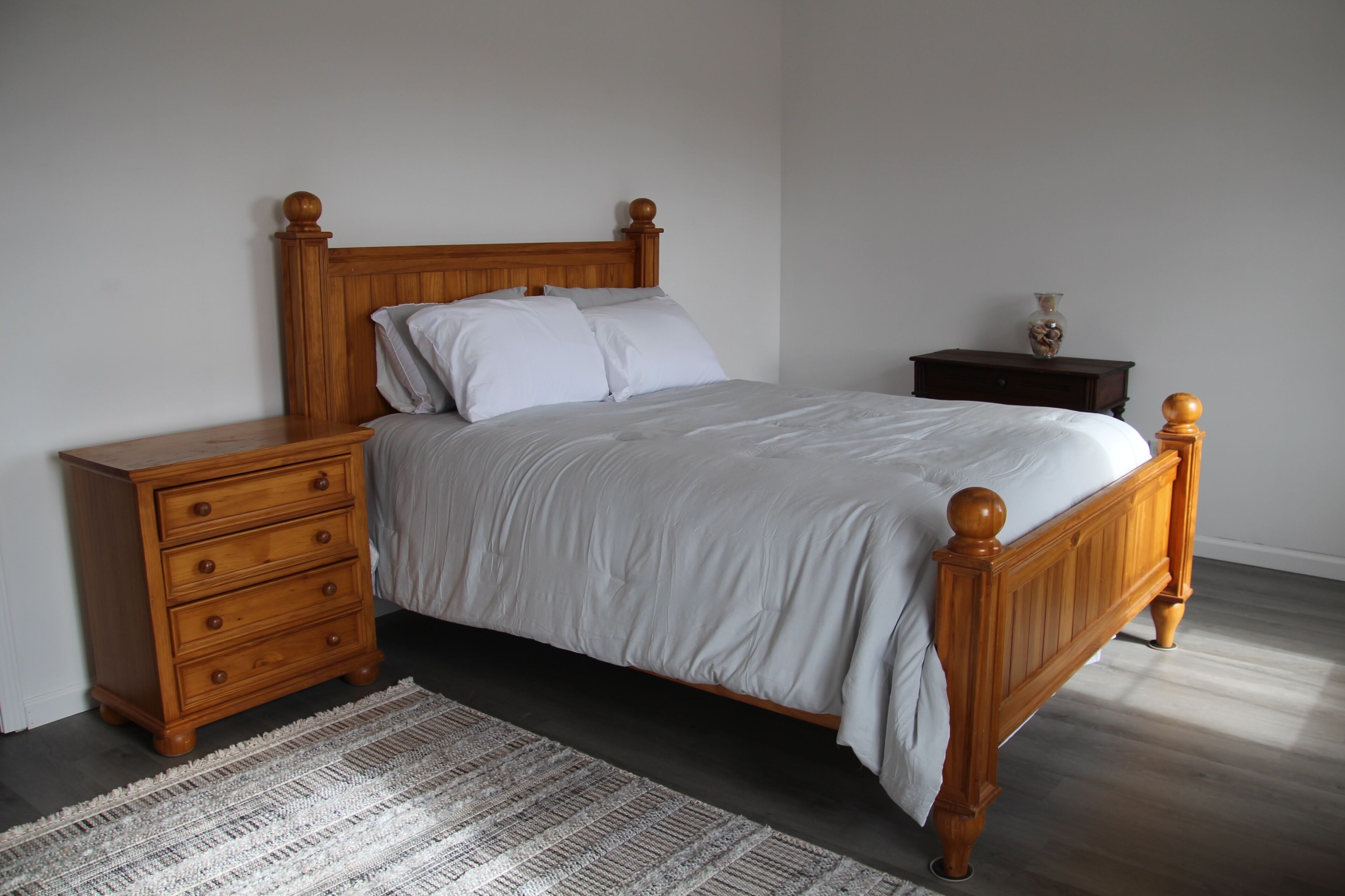 Un dormitorio con una cama de madera, una mesita de noche con cajones, una pequeña mesa con un florero y una alfombra estampada.