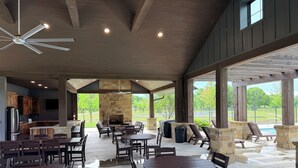A covered patio area with multiple tables and chairs, a stone fireplace, and a view of a swimming pool.