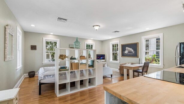 Una sala de estar moderna con piso de madera, muebles blancos, una estantería, un comedor y una cocina con encimera negra.