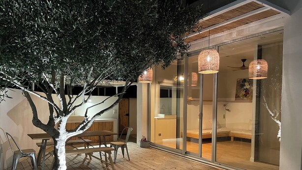 Un espace repas extérieur avec une table et des chaises en bois, un arbre et une porte vitrée donnant sur une chambre à coucher équipée d’un lit et d’un ventilateur de plafond.