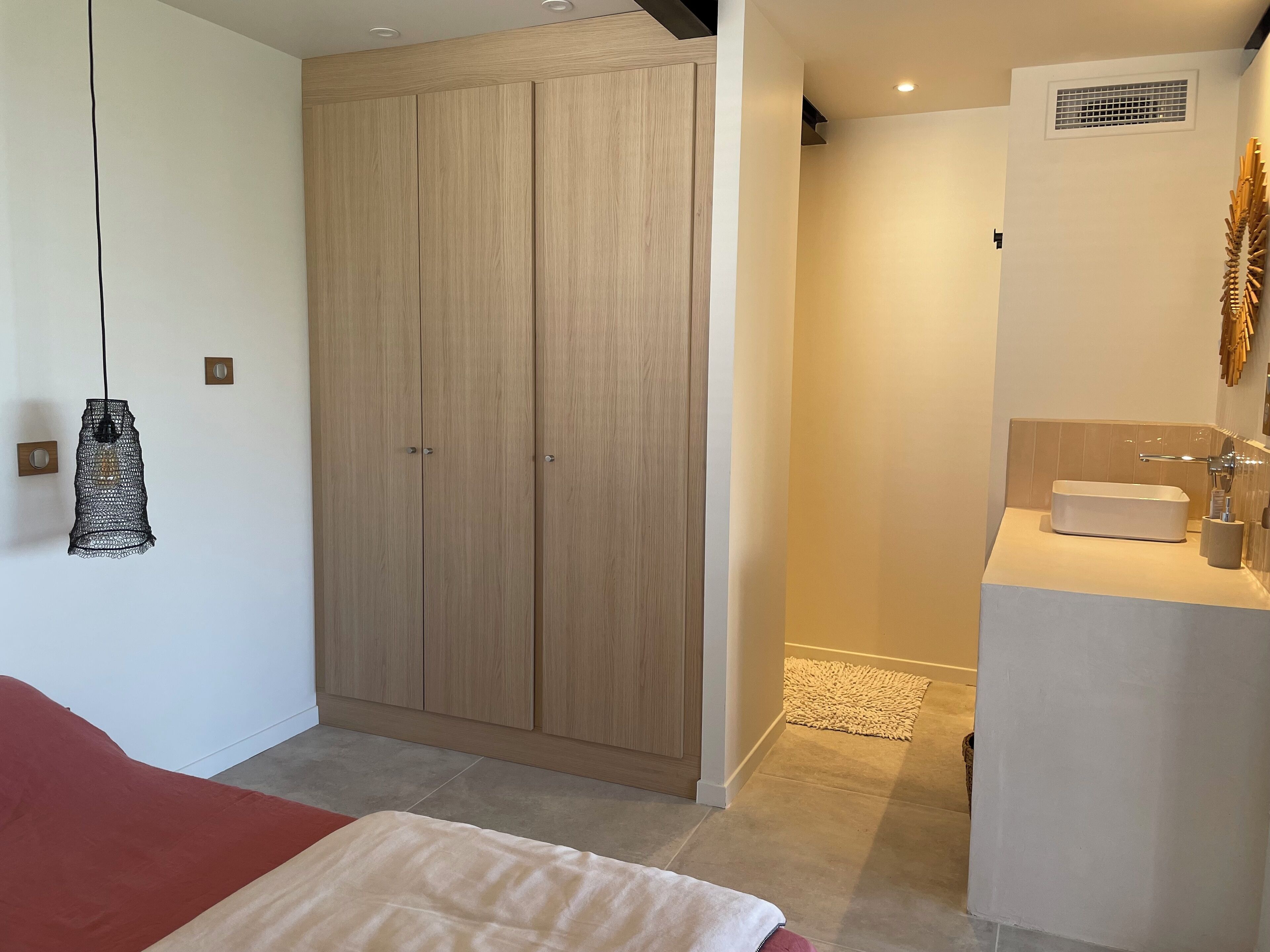 Une chambre à coucher comprenant un lit, une armoire, un luminaire suspendu et une salle de bain avec un lavabo et un miroir.