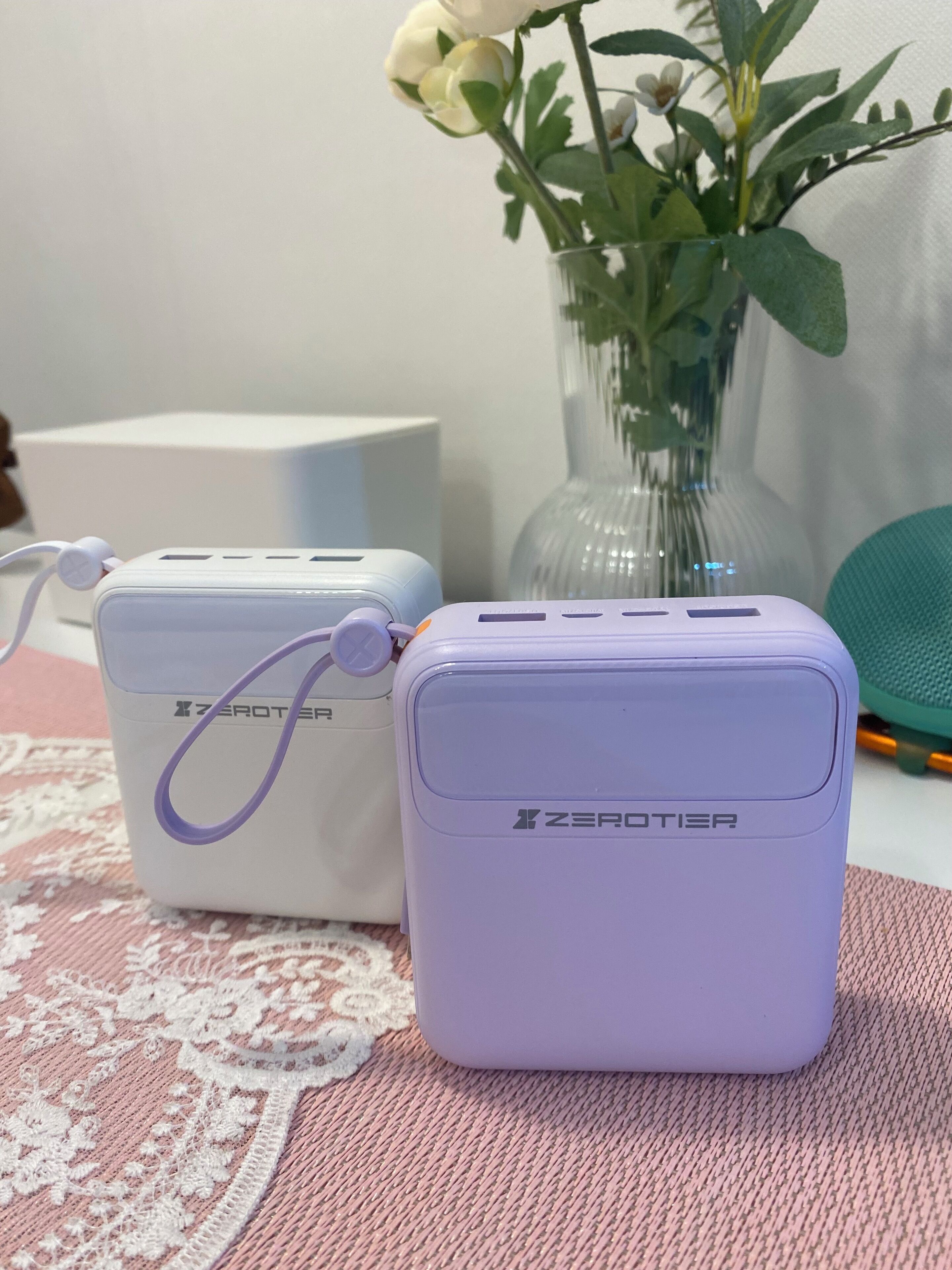 Two ZeroTier portable chargers on a pink textured surface with a white vase and a green chair in the background.