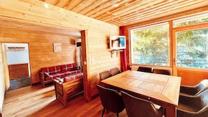 A wooden cabin interior with a dining table, chairs, a red plaid sofa, a television on a wall, and a large window offering a view of trees. - Chalet Alpe One 1850 M - Alpe d'Huez (Huez)