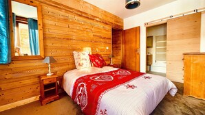 A bedroom with wooden walls, a bed with a red and white blanket, a nightstand with a lamp, and a mirror. - Chalet Alpe One 1850 M - Alpe d'Huez (Huez)