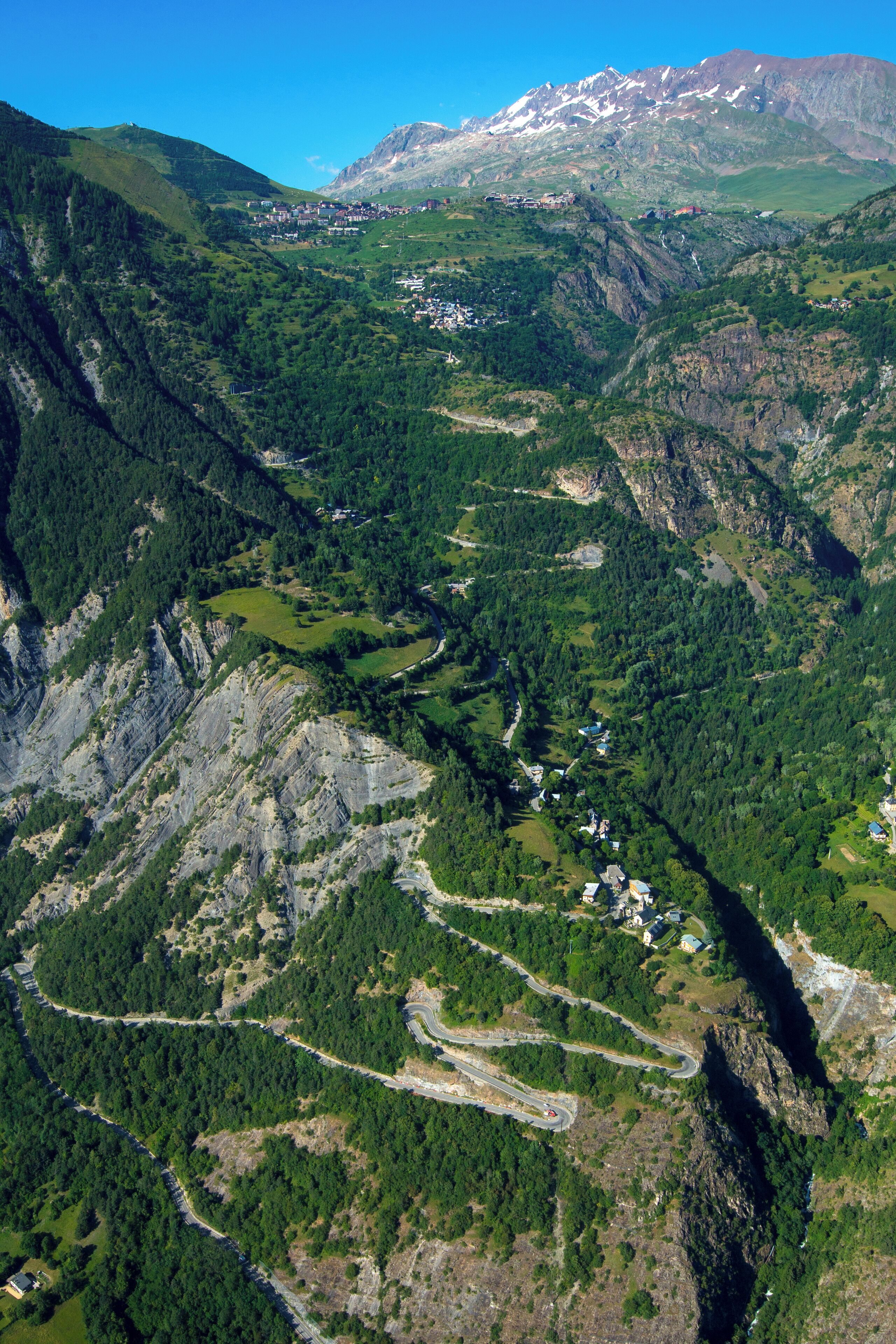 A mountainous landscape with a winding road, dense forests, and a small settlement with buildings.