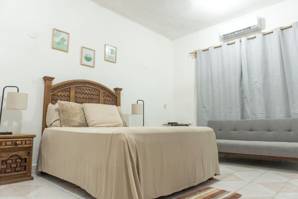 Un dormitorio con cama, mesitas de noche, un sofá gris y una ventana con cortinas.