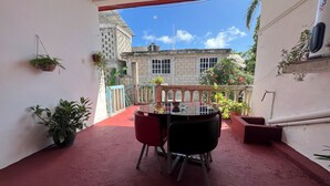 Un balcón con una mesa y sillas, plantas en macetas y vistas a un edificio con baranda azul y suelo rojo.