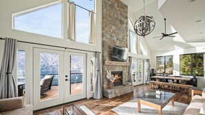 A spacious living room with a fireplace, large windows, and a view of a lake. - NEW! Gorgeous lake home w/ private pool, hot tub, lake access with dock & views! (Manson)