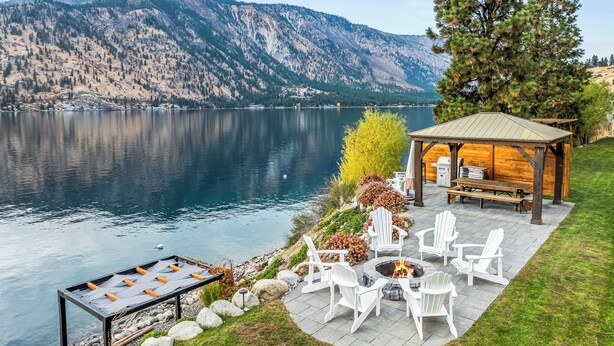 A lakeside patio with a fire pit, Adirondack chairs, and a wooden gazebo overlooking a mountain lake. - NEW! Gorgeous lake home w/ private pool, hot tub, lake access with dock & views! (Manson)