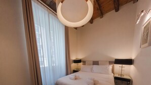A hotel room with a bed, two bedside tables, a nightstand, a hanging light fixture, and a window with curtains. - Vatican Mascherino suite (MAS-2) (Roma)