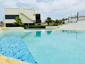 A swimming pool with a tiled edge, surrounded by a modern building with a flat roof and a fence. - Apartment with large terrace in Punta Prima, (VC)