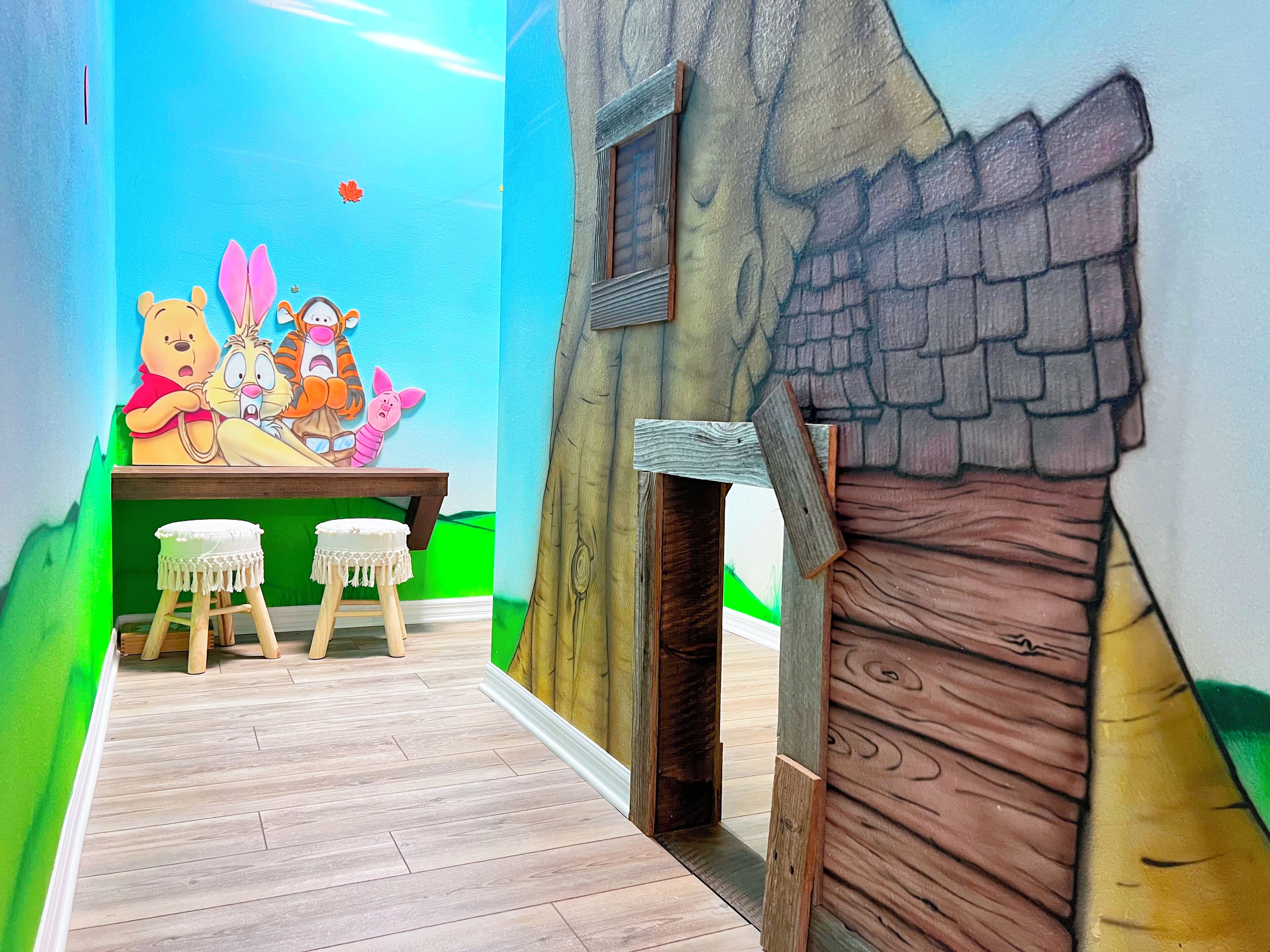 A children's playroom with a mural of Winnie the Pooh and friends, two stools, and a wooden playhouse structure.