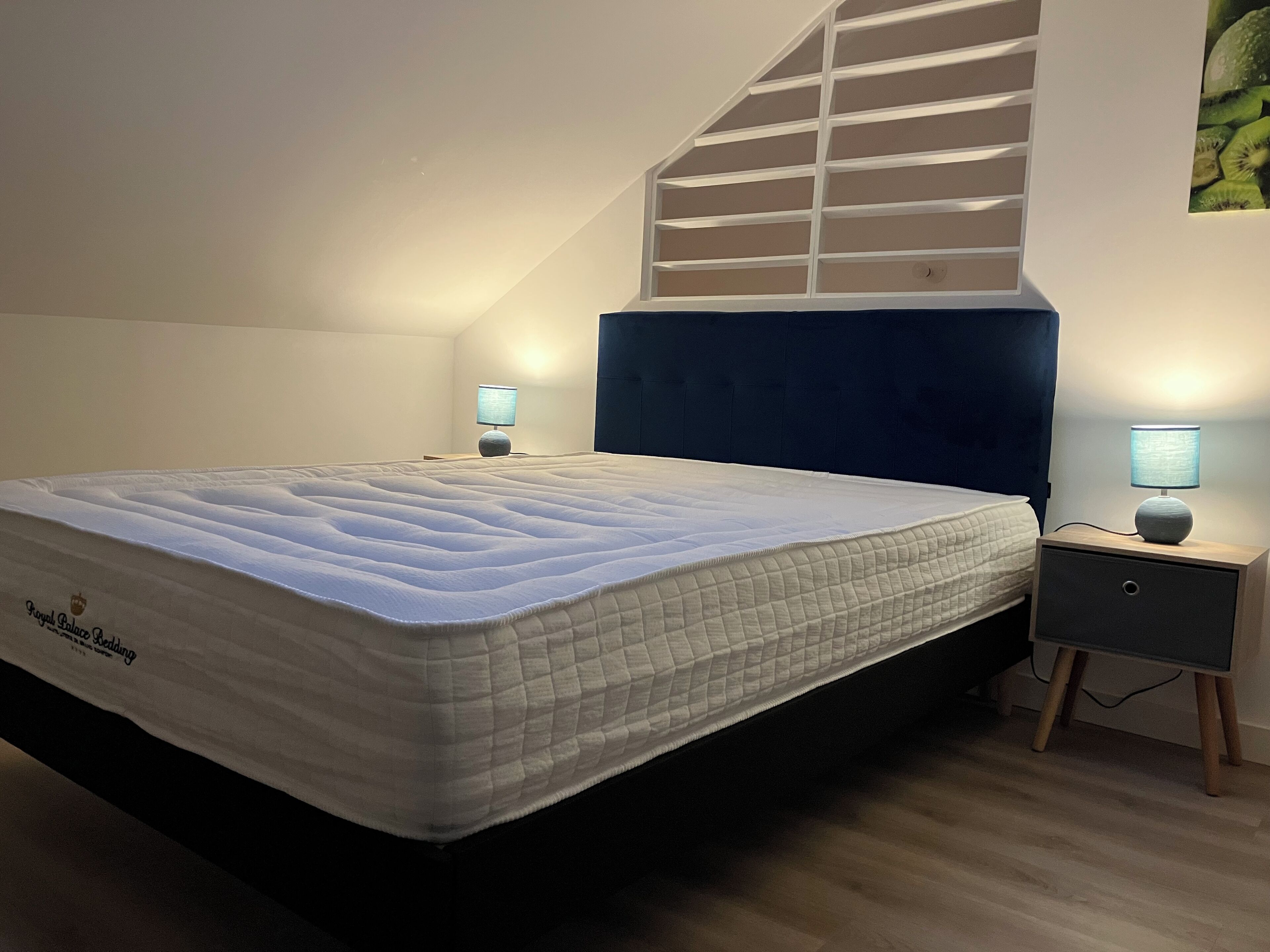 Ein Schlafzimmer mit einem großen Bett, zwei Nachttischen mit Lampen, einem dunklen Kopfteil und einer schrägen Decke mit einem Fenster.