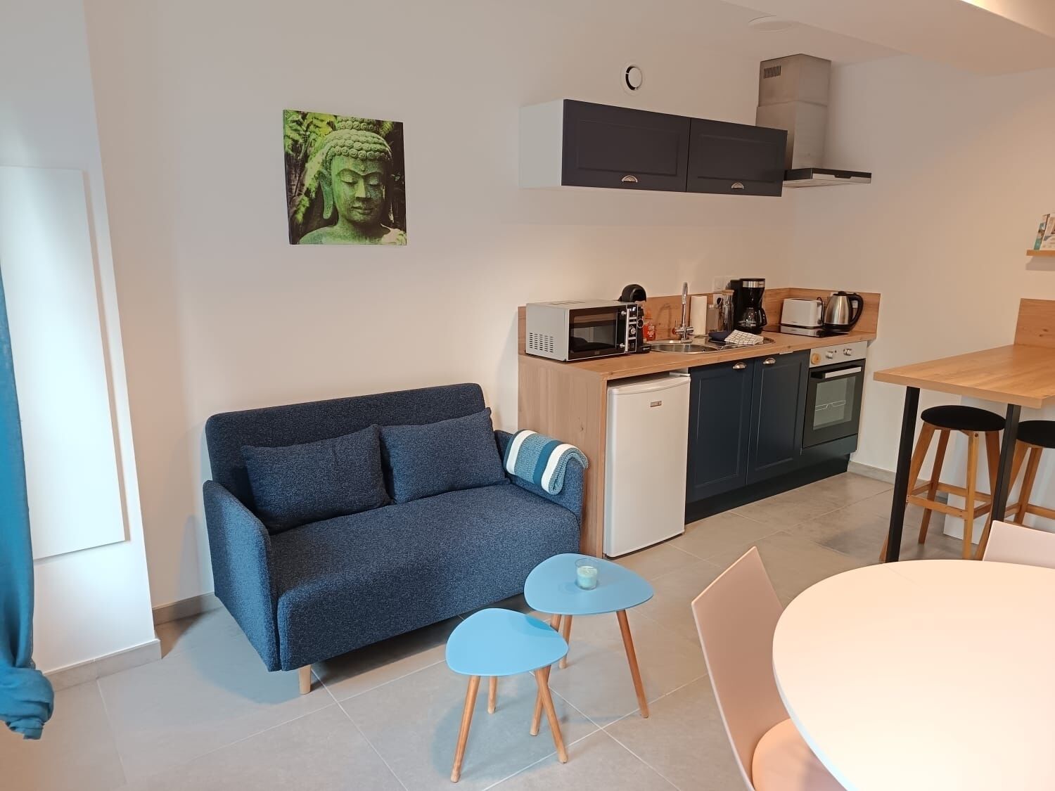 Een compacte woonruimte met een bank, een kleine keuken met een magnetron, koffiezetapparaat en koelkast. Er is een eettafel met stoelen en een klein blauw tafeltje.
