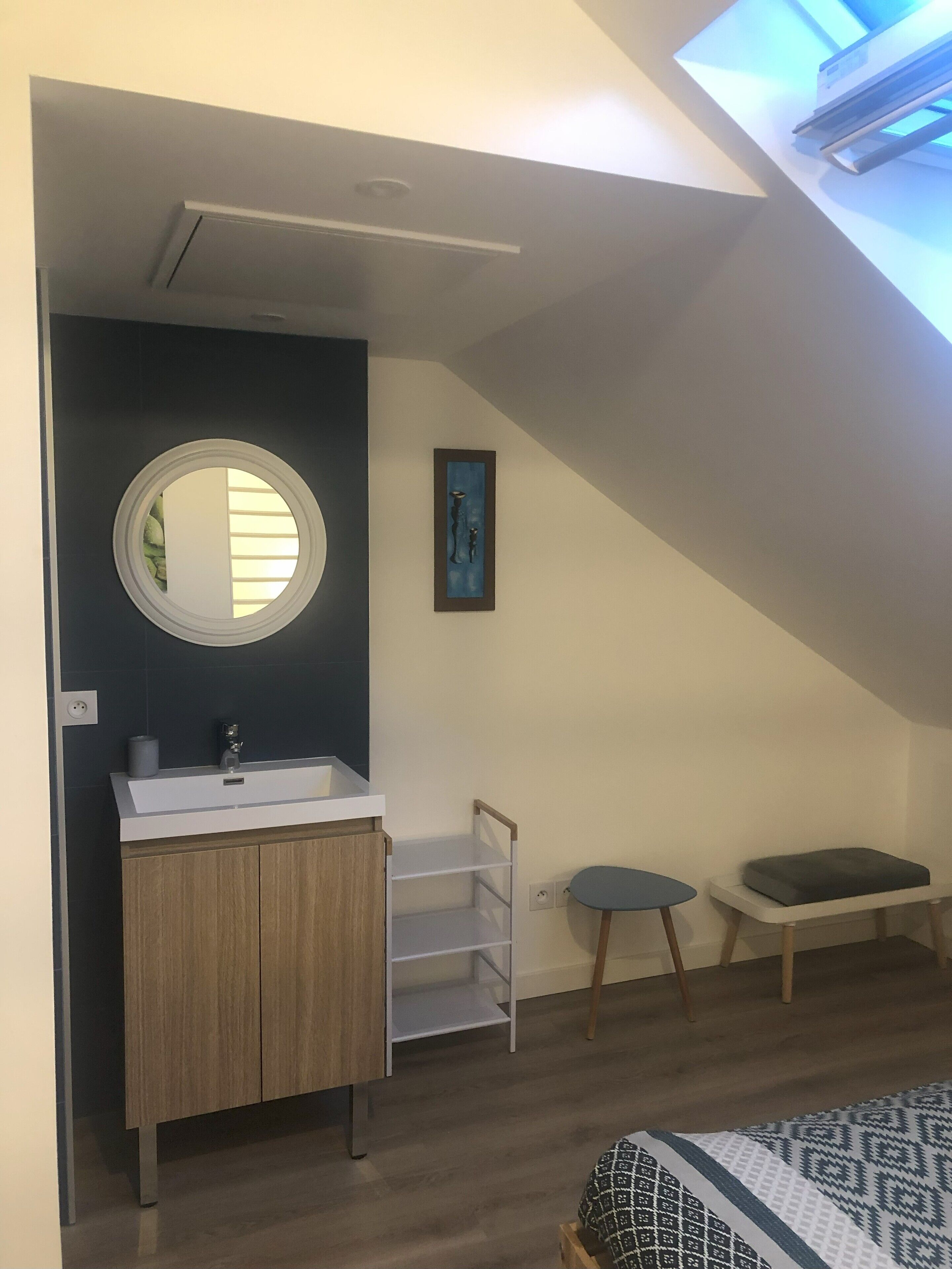 Ein Schlafzimmer mit Dachfenster, einem runden Spiegel, einem Holzschrank, einem kleinen Tisch und einem Bett mit gemusterten Betttextilien.