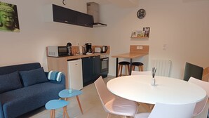 A compact living space with a sofa, a small dining table with chairs, a kitchen area with appliances, and a clock on the wall.