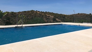 Une piscine dotée d’une plage en béton, entourée d’une clôture en métal, et offrant une vue sur un paysage vallonné en arrière-plan.