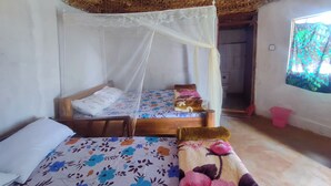 A room with a bed under a mosquito net, another bed with a floral bedspread, a wooden headboard, a window with a view, and a woven ceiling.
