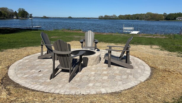 A circular outdoor seating area with Adirondack chairs and a table, overlooking a lake with a dock and boat in the background. - Modern Lake Home: Family Fun & Memorable Getaway! (Beaver Dam)