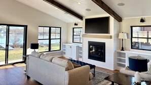 A spacious living room with a high ceiling, large windows, a fireplace, and built-in shelving. - Modern Lake Home: Family Fun & Memorable Getaway! (Beaver Dam)