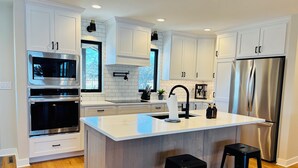 A modern kitchen with white cabinetry, stainless steel appliances, and a central island with stools. - Modern Lake Home: Family Fun & Memorable Getaway! (Beaver Dam)