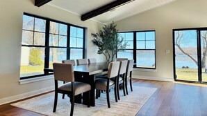 A dining area with a large table and chairs, large windows with a view of a body of water, and a wooden ceiling. - Modern Lake Home: Family Fun & Memorable Getaway! (Beaver Dam)