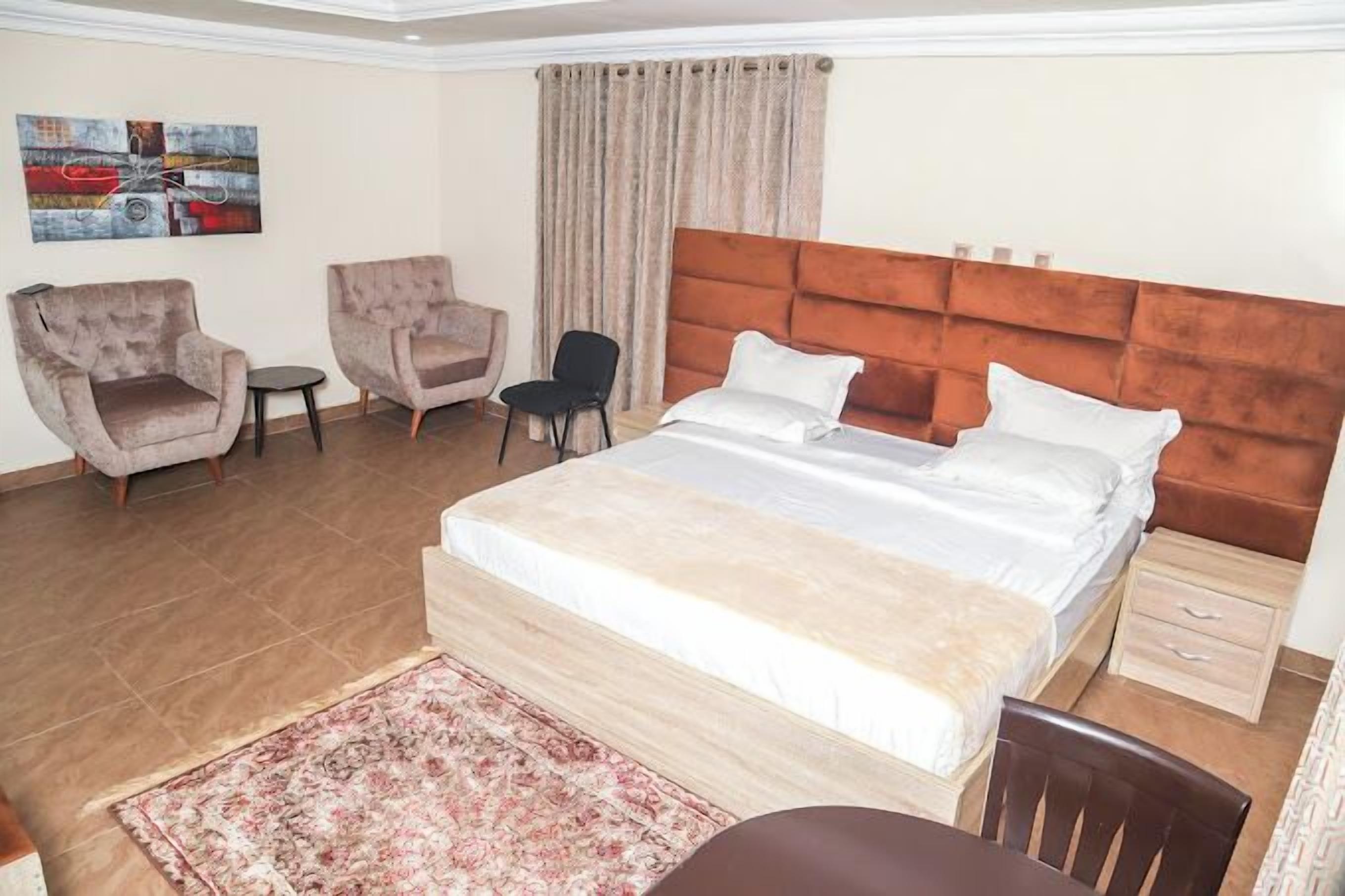 A hotel room with a large bed, two armchairs, a small table, a chair, and a nightstand.