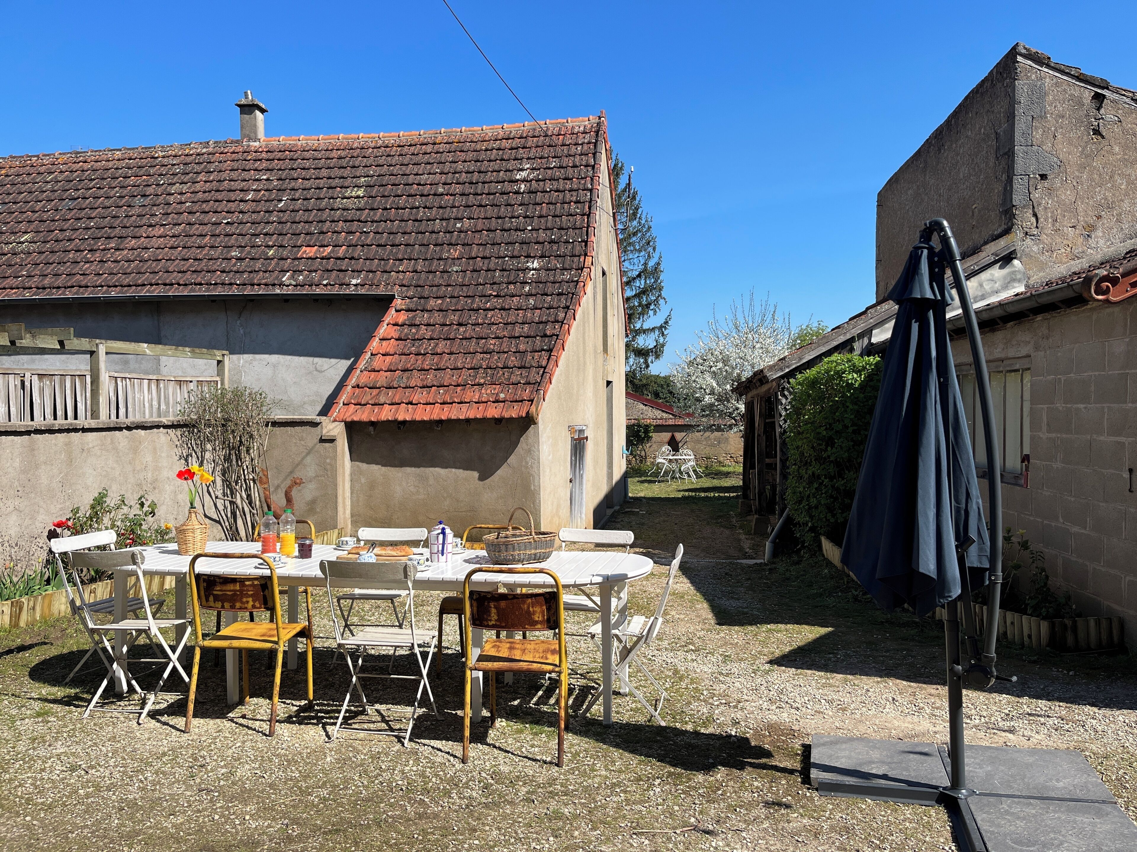 Un coin repas extérieur rustique, avec une table et des chaises, un panier en osier et une bouteille de jus d’orange.