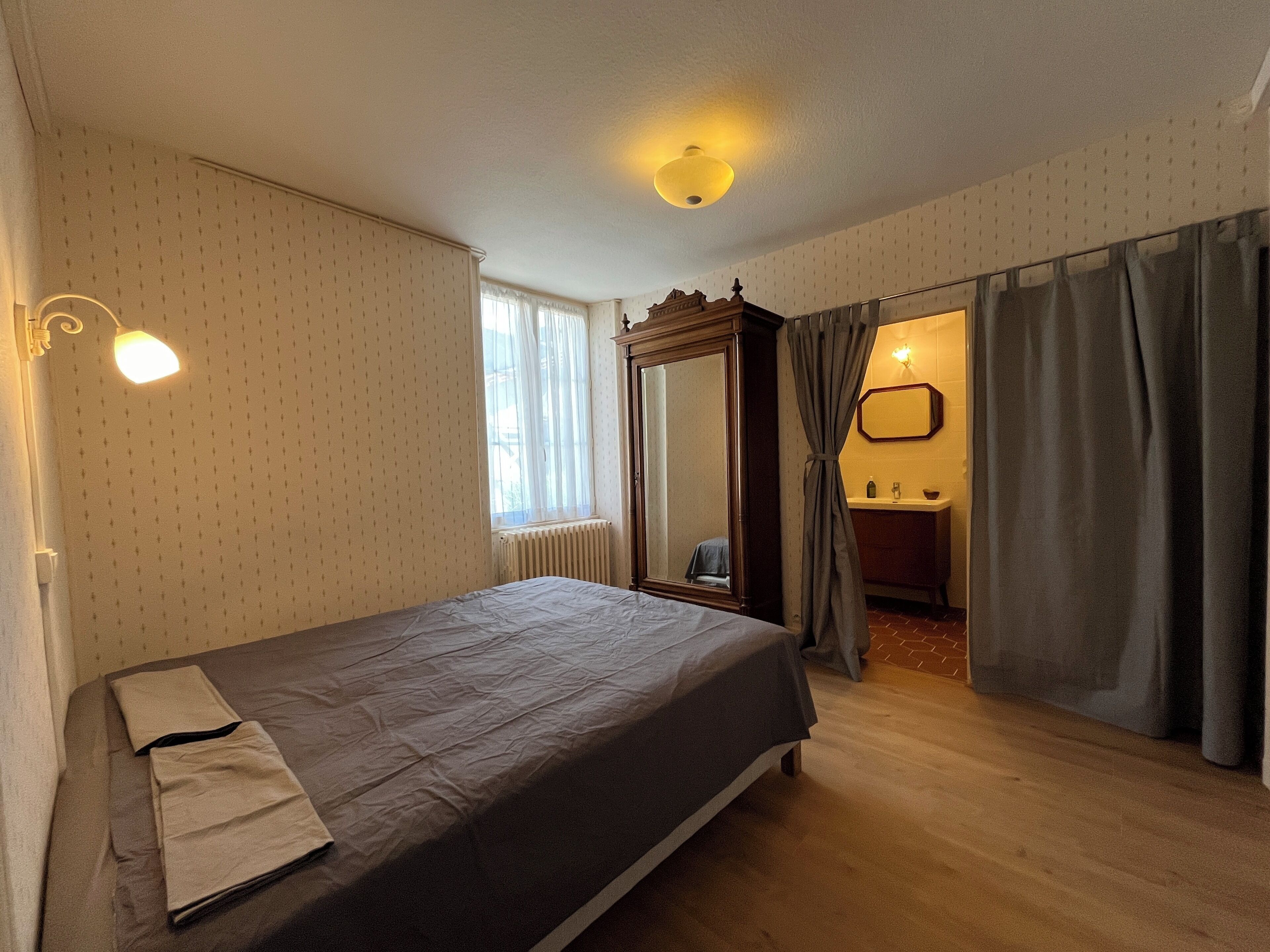 Ein Schlafzimmer mit Bett, Fenster, Heizkörper, Spiegel und einem durchgehenden Türrahmen erkennbaren Badezimmer.