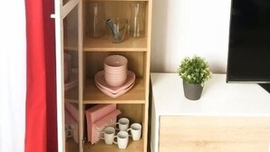 Ett rum med en hylla i trä där det står glasvaror, rosa glas i form av flamingos och tallrikar. En liten krukväxt står på ett bord bredvid en platt-TV.