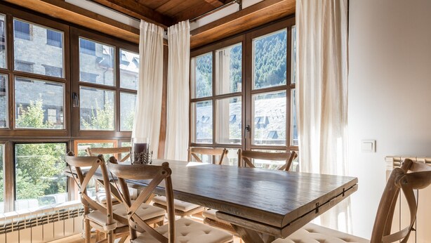 Un comedor con mesa y sillas de madera, ventanas grandes con cortinas y vistas a edificios y árboles.